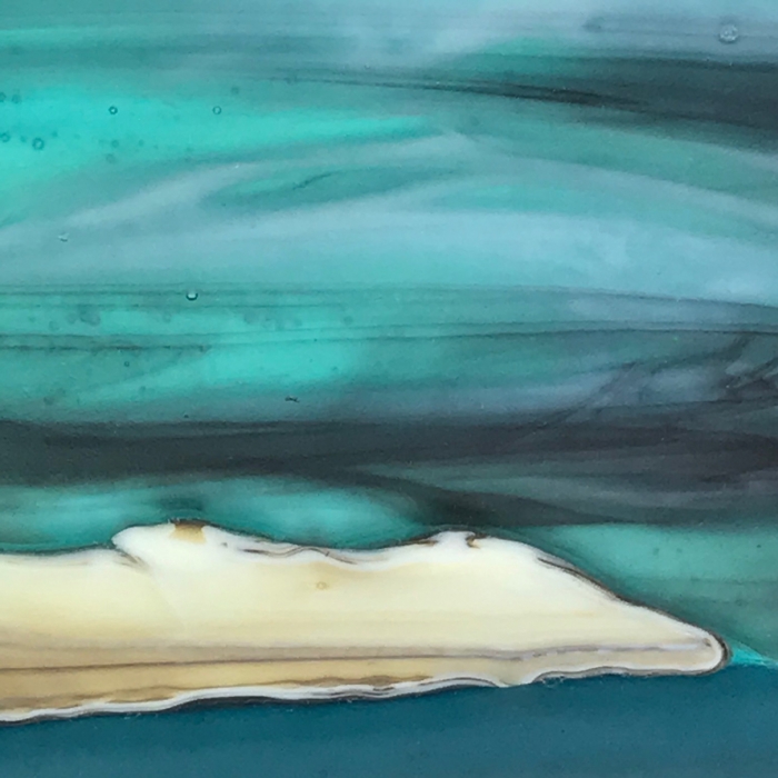 Glass Wall Art, welsh seascape. Aquamarine sky with moody clouds. Steel blue sea. Headland, foreground with french vanilla/browns/teals glass frits to fore.Detail.