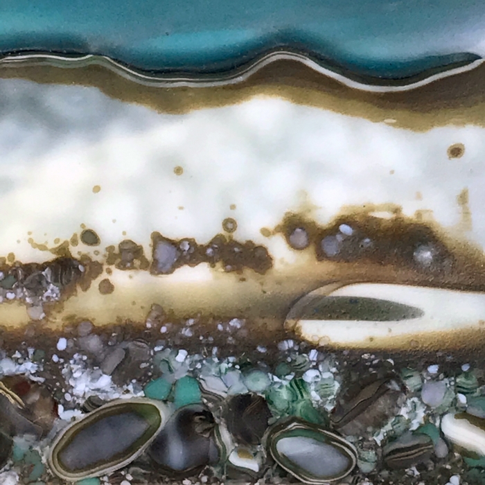 Glass Wall Art, welsh seascape. Aquamarine sky with moody clouds. Steel blue sea. Headland, foreground with french vanilla/browns/teals glass frits to fore. Detail.